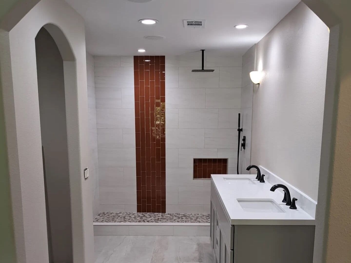 After: the same arched entry opening onto white large-format tile, a rust glass vertical accent, pebble shower floor and a new double vanity