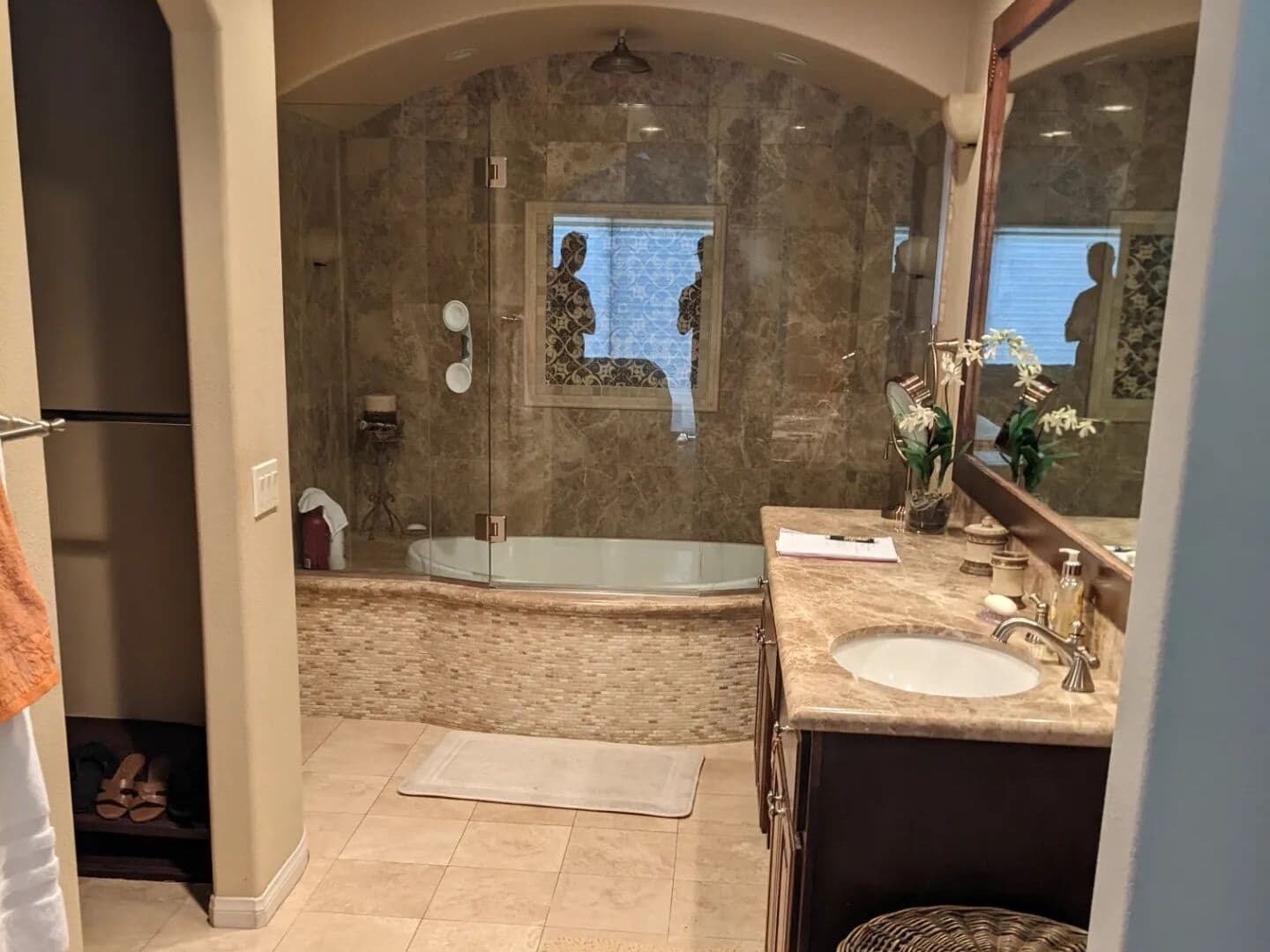 Before: dated master bath seen through the arched entry, with a brown marble shower, corner tub and tan tile floor