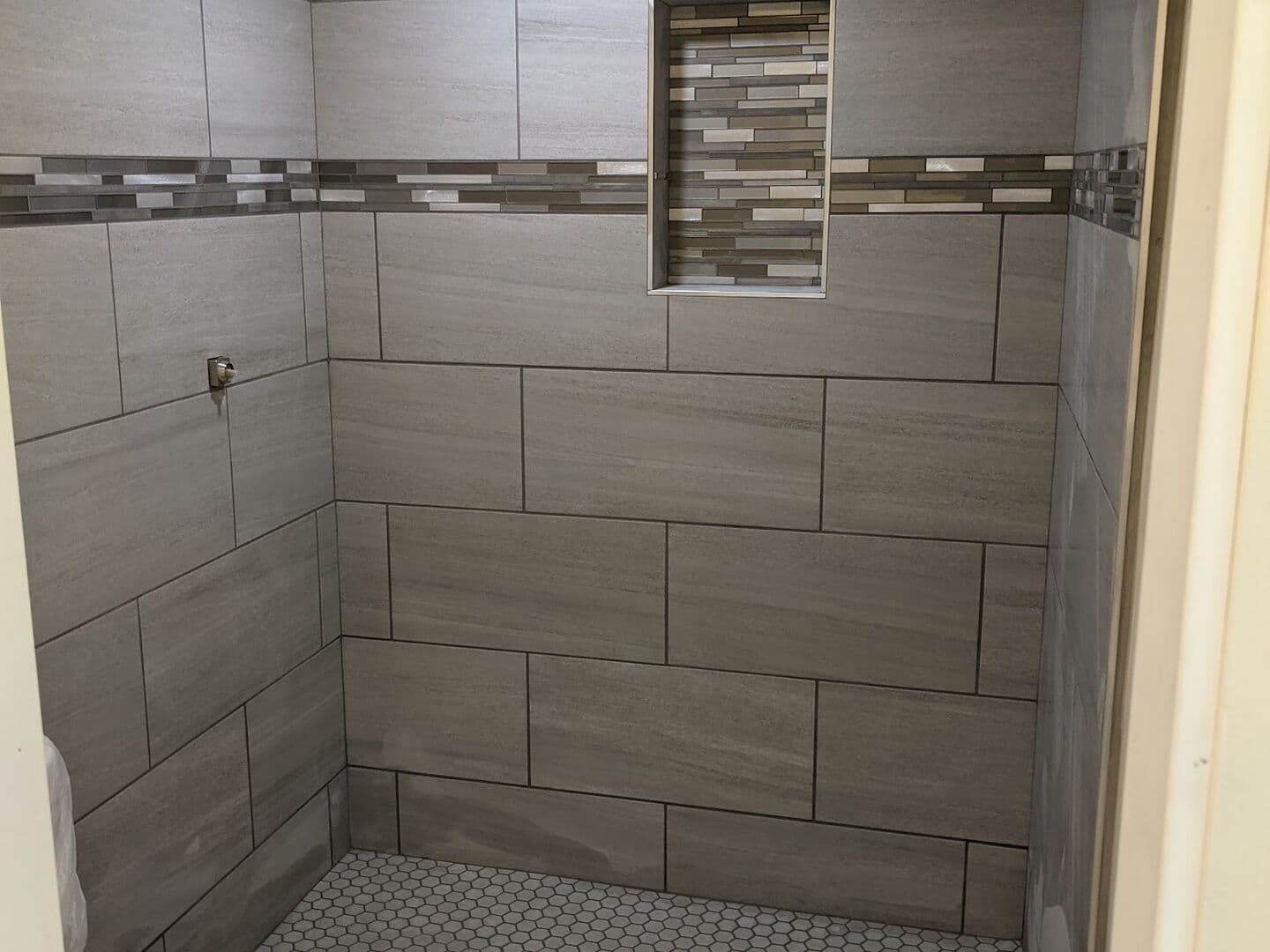 Walk-in shower in grey linear porcelain with a glass mosaic band, recessed niche and marble hex floor