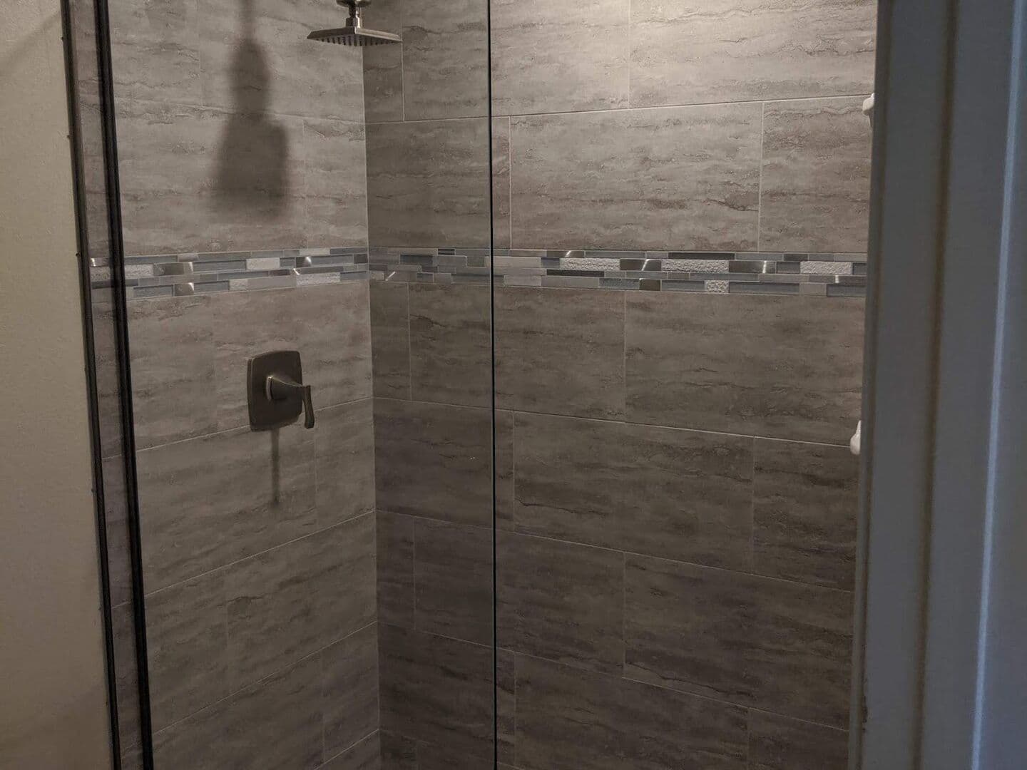 Walk-in shower in grey wood-look tile with a glass mosaic band, pebble floor and frameless glass panel