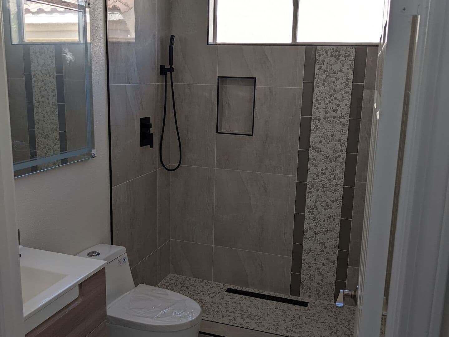 Remodeled bathroom with a floating wood vanity, wall-hung toilet and tiled walk-in shower with a vertical accent stripe