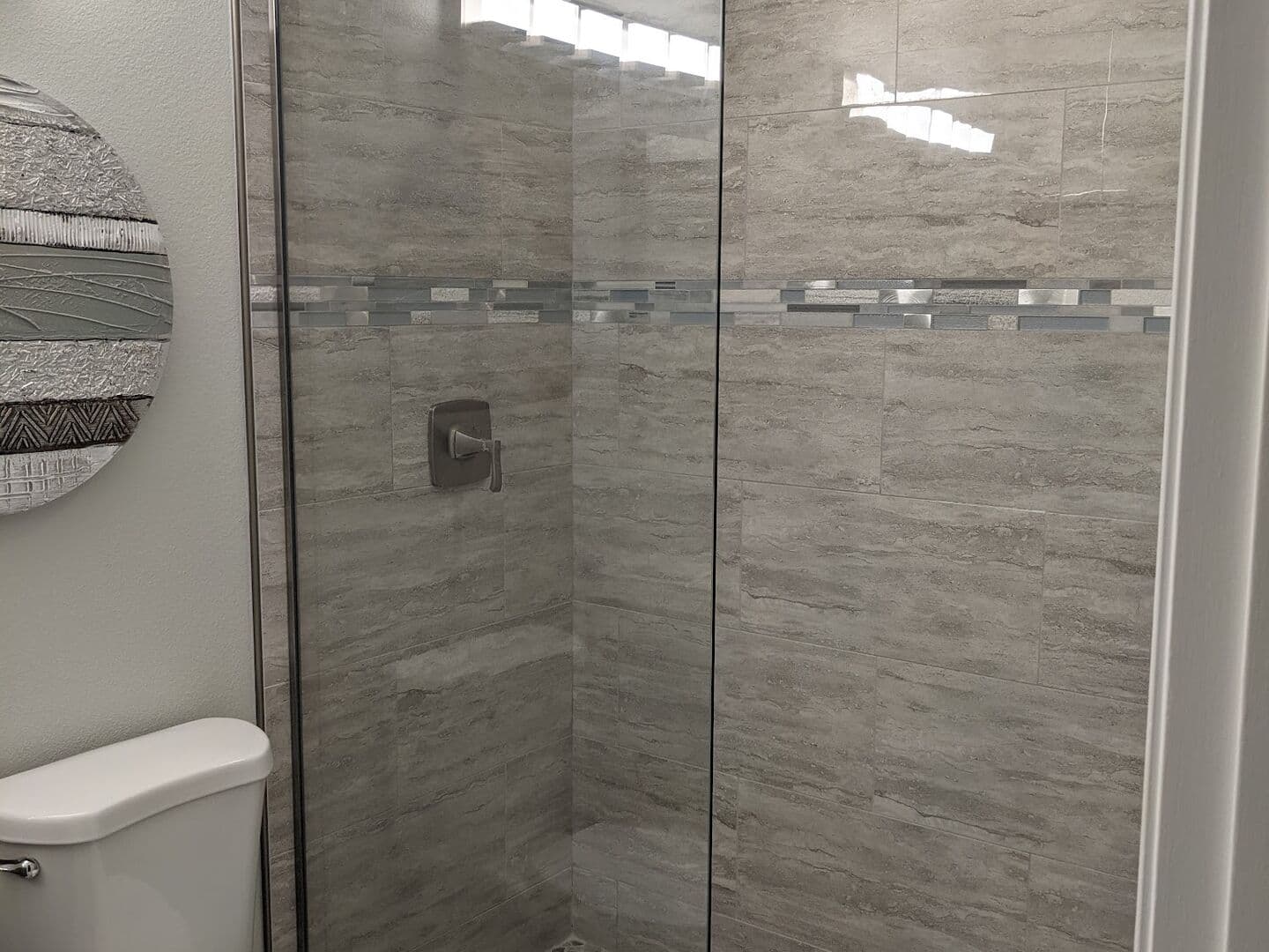 After: tub removed and the alcove rebuilt as a tiled walk-in shower with a glass mosaic band, pebble floor and frameless glass panel